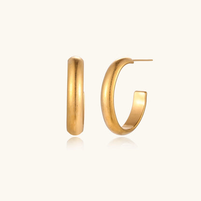 Chunky tubular hoop earrings in warm 14K gold-plated finish with smooth polished surface on cream background.